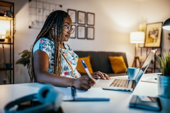 A student writes in a notebook while looking at a laptop with her living room in the background. 