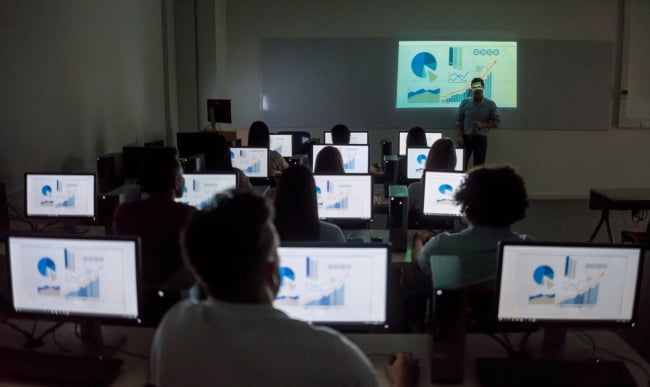 A group of students sits in front of computers, each showing charts and graphs on the screen. They are all facing toward the front of the room with their backs turned toward the camera.