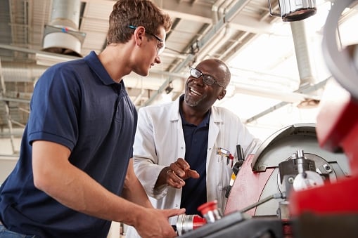 Engineer talking to a younger male apprentice over machinery in a factory/research setting. 