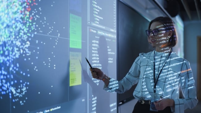 A woman points toward a screen filled with electronic blue dots. She also has electronic text projected onto her