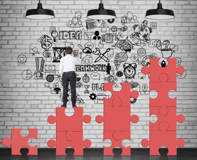 Rear view of businessman standing on red puzzle pieces and drawing business graffiti on brick wall, with the words Teamwork and Idea visible.