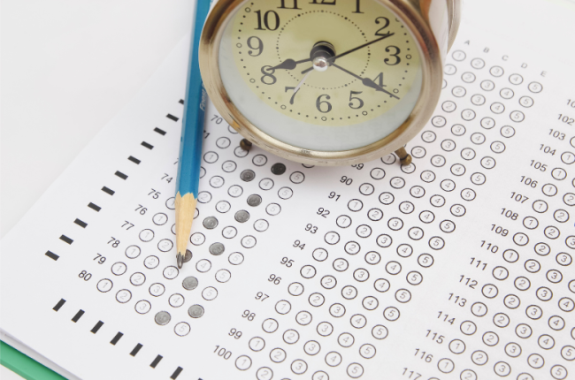 A standardized test answer sheet with bubbles filled in. A pencil and a small circular clock sit atop the sheet.