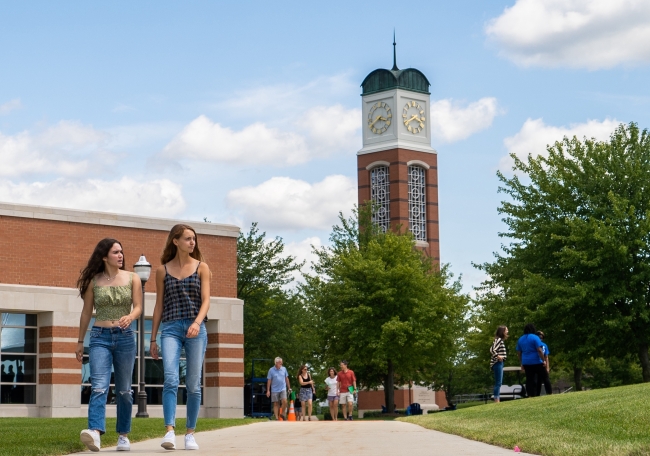 Two girls walk on a sidewalk with a college campus behind them. 