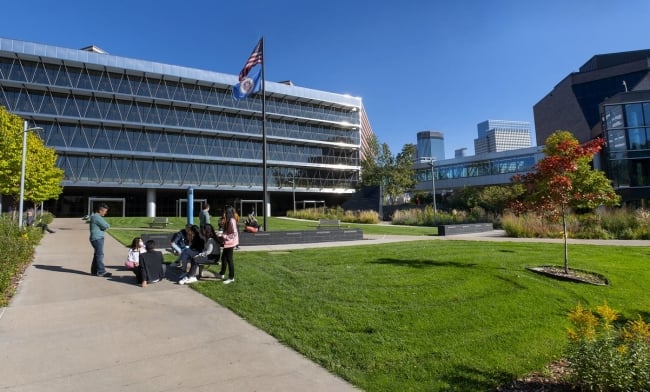 A group of people gather on a sidewalk next to a green lawn under a waving Minnesota state flag