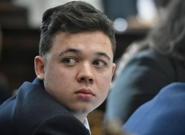 A close up of Kyle Rittenhouse, a young white man with brown hair, glancing behind himself while in a courtroom.