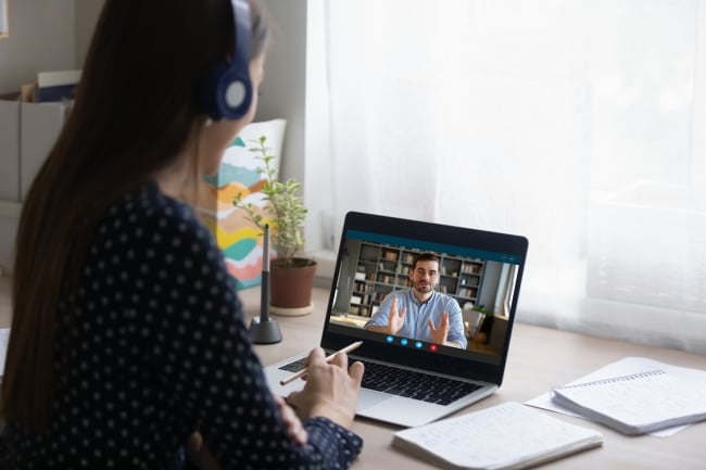 A woman wearing headphones faces a laptop. A man is on the screen of the laptop teaching. 