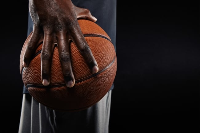 A close-up photo of a Black athlete’s hand holding a basketball, against a black background..