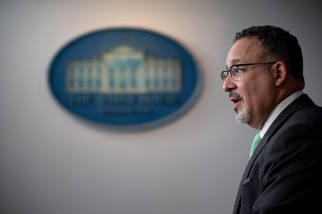 Education Secretary Miguel Cardona stands at a podium. A blurred-out image of the White House seal is in the background.