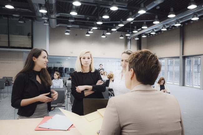 Three students stand before a teacher, one with arms crossed as if annoyed about something