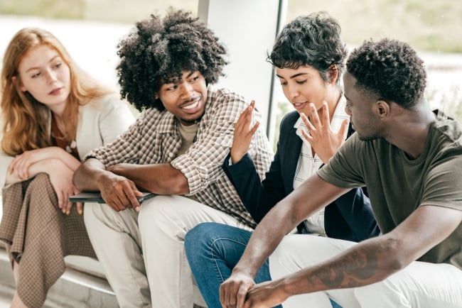 A group of four young adults talk animatedly next to a large window on campus.