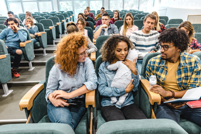 A young mother holds her baby in a lecture hall, with other students sitting around her.