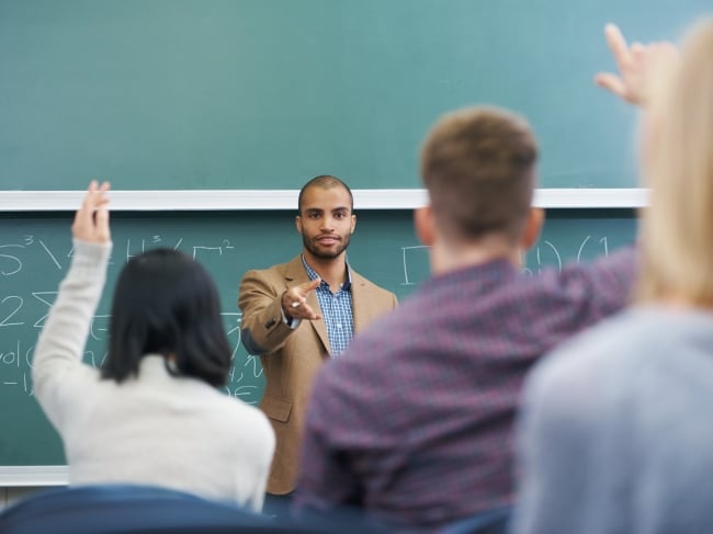 A young male teacher points at two students in the foreground who have their hands raised. Behind the instructor is a chalkboard with math equations