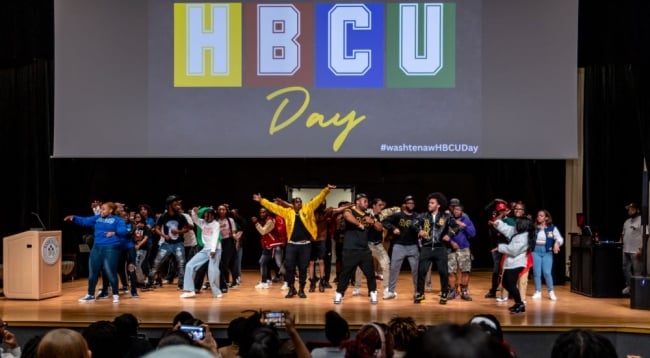 A group of students dance on a stage with a screen that reads “HBCU Day” in the background. 