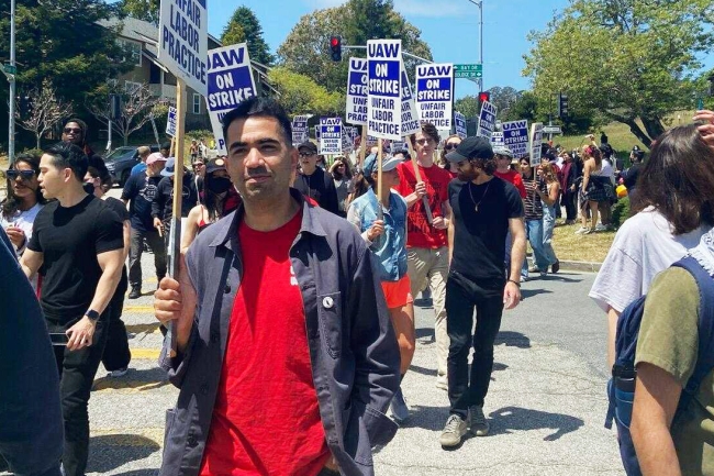 A photograph of people marching and holding signs that say “UAW On Strike Unfair Labor Practice.”