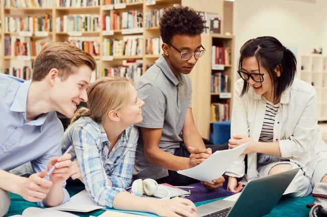 Four students sit around a desk, looking at papers and laptops.