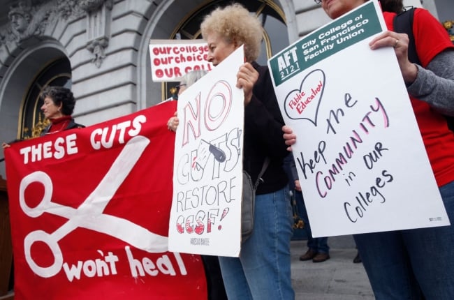 Three women stand with signs. One red sign features scissors and reads, “These cuts won’t heal.”