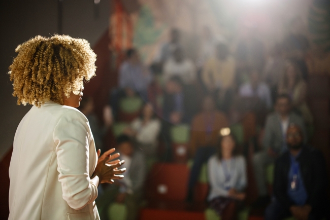 Woman stands with her back to camera facing large audience and bright light