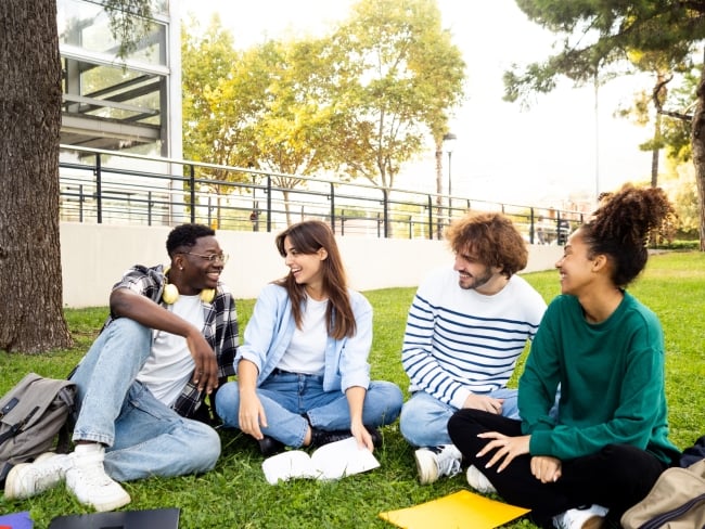 Group of diverse college student friends laughing and chatting sitting on the grass on campus