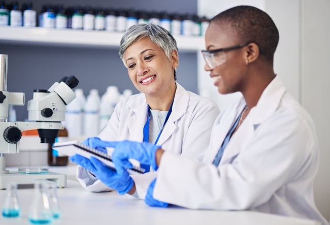 Older scientist helping younger scientist. Both are women.