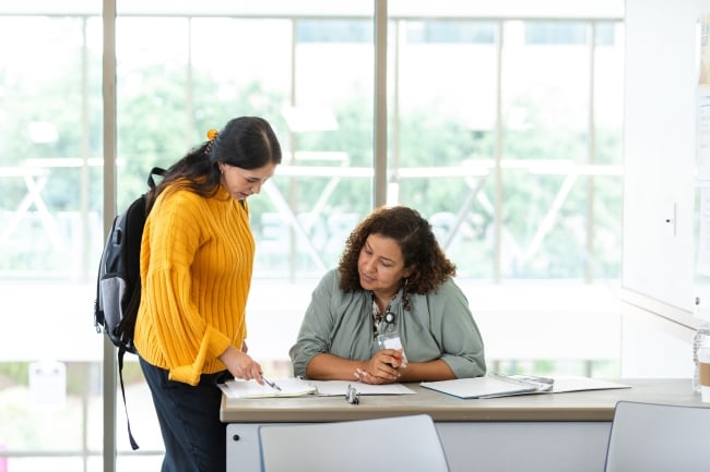 A young adult female student consults a mature adult teacher's before leaving class.