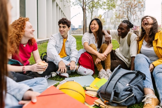 Group of diverse college students sitting outside on grassy campus area laughing and talking