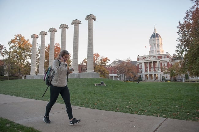 A student walks down a path on a college campus in front of a row of columns