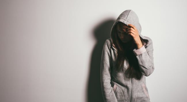 A college student stands in a poorly lit room with hood on her head and hand over her face, looking embarrassed.
