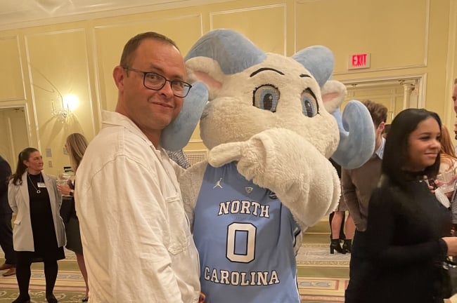 A man in glasses with the University of North Carolina ram mascot in its Carolina blue basketball uniform