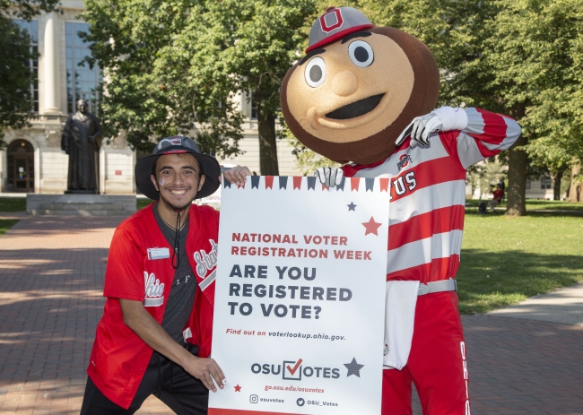 A student standing beside Ohio State University mascot Brutus Buckeye holding a sign asking students if they are registered to vote.