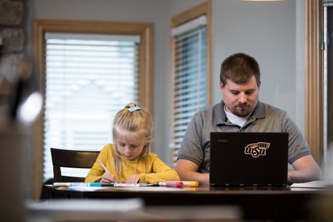 An adult student works at a laptop while his young daughter colors next to him