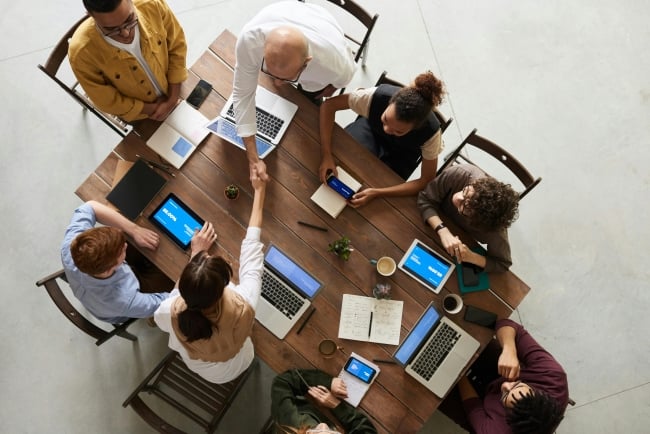 Aerial photo of a group of professional people gathered around a table with laptops and phones, some shaking hands.