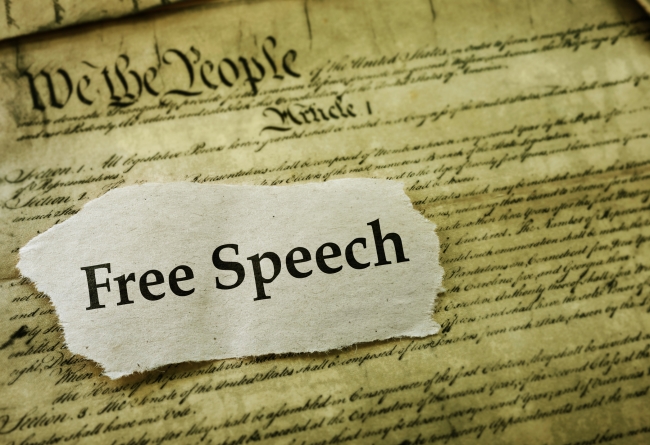 A cutout of a piece of paper with the words "Free Speech" laid atop a sepia-toned copy of the U.S. Constitution.