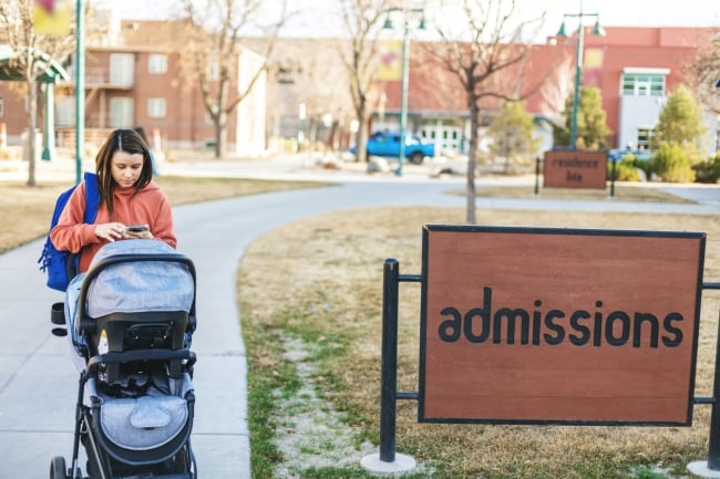 A woman pushes a baby stroller on a college campus next to a sign that says "admissions." She is texting on her phone as she walks