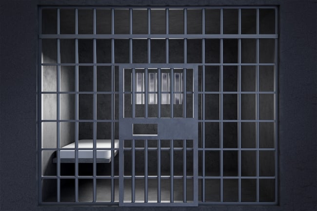 A illustration of a jail cell, taken from outside the bars, with a single bed visible.