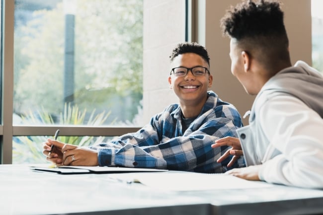 Two young Black men sit at a desk and chat, smiling