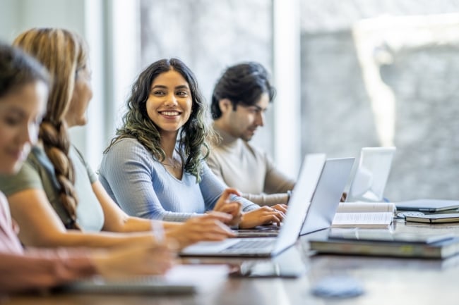 A small group of four mature adult students sit together at a long desk as they study with one another. They have textbooks and laptops out in front of them as they take notes and talk.