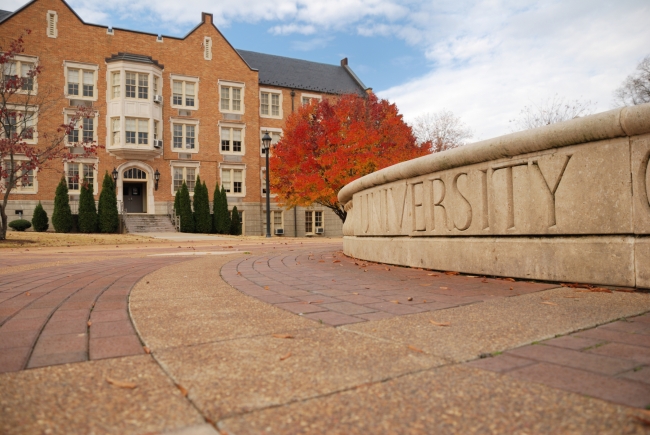 A stock photo of a university building next to a sign that reads "university," next to a tree in autumn with red leaves.