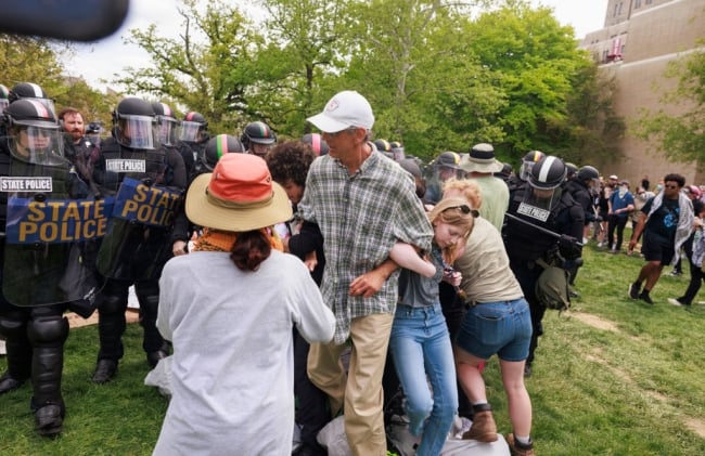 A photograph of police in riot gear, with the words "state police" on their shields, approaching a group of people huddled together, holding on to one another, on a lawn at Indiana University at Bloomington.