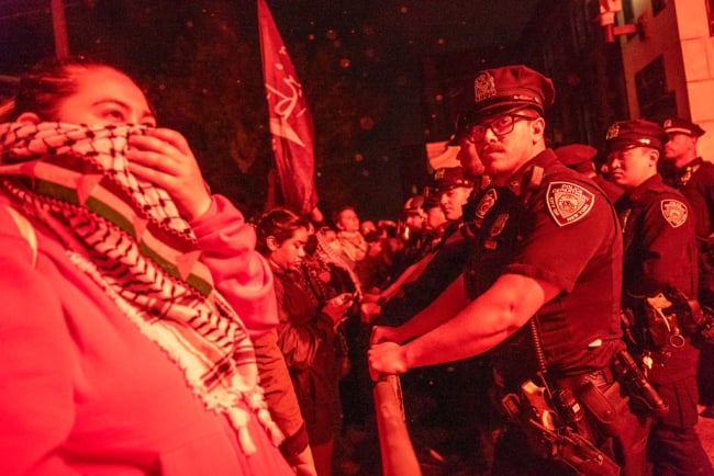 In this red-toned image, pro-Palestinian protesters stand face-to-face in a confrontation with police at the City College of New York on April 30. In the foreground, a protester holds a keffiyeh to her face as a mask while a male police officer looks on.