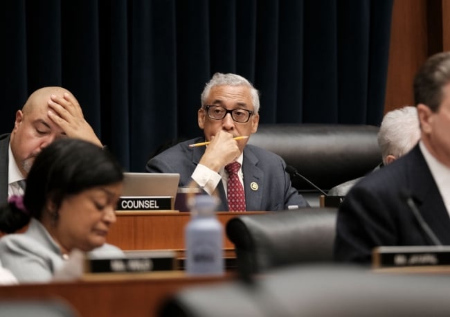 A light-skinned Black man with gray hair wearing a suit and tie sits at a desk