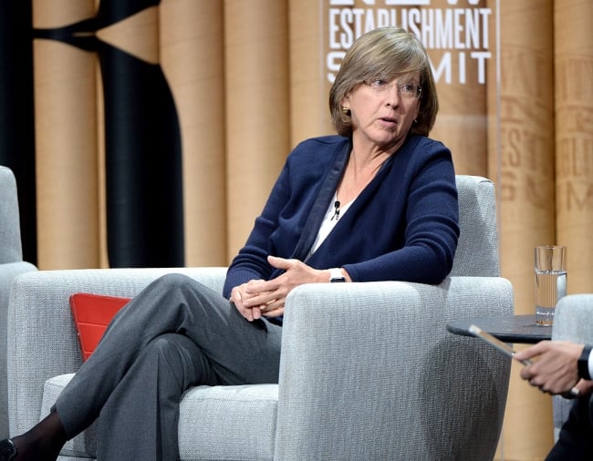 A woman with glasses wearing in a blue sweater sits onstage, leaning on the arm of her chair as she speaks to someone next to her.