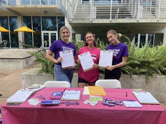 Three young woman stand behind a table holding voter registration forms.