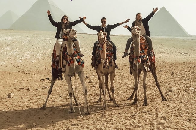 Three students sit on camels in a desert.