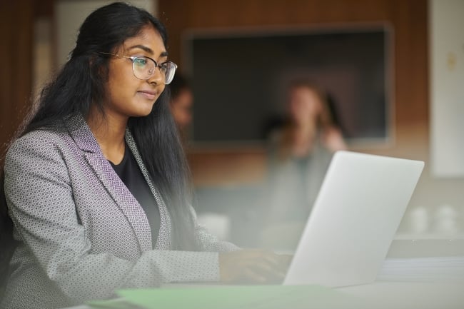 College student wearing a blazer and glasses works at a laptop.