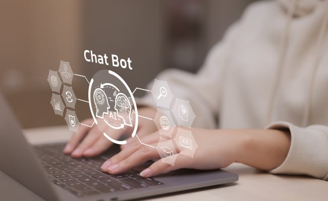 A close-up of a woman's hands resting on a laptop keyboard. An illustrated icon that reads "chat bot" hovers above the computer and the woman's hands.
