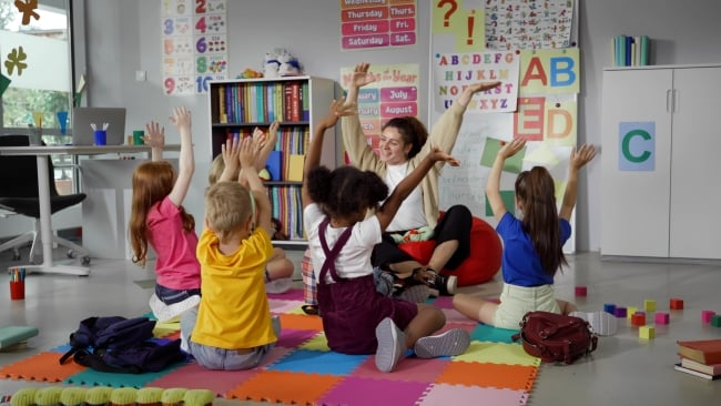 A teacher sits on the floor with a group of young students, all with their hands in the air. 