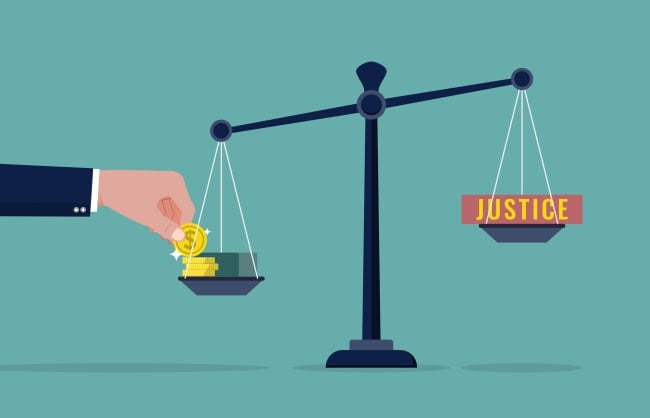 Hand placing money on one side of a scale weighing down the word "justice" on the other side of the scale