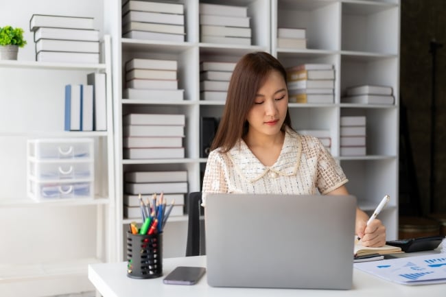 Asian woman sits in front of a bookcase and at a desk with a computer and charts 