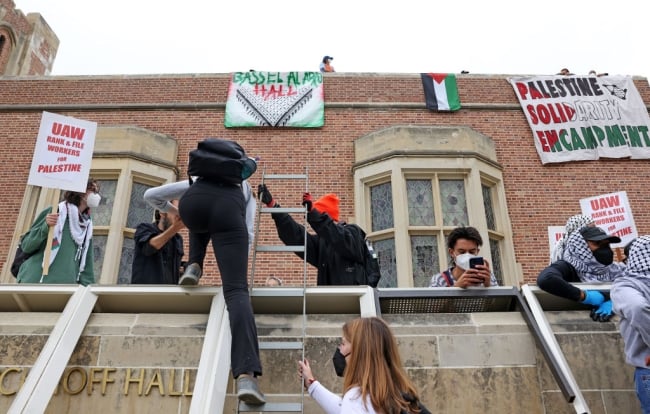 Pro-Palestinian protestors trying to set up a solidarity encampment at the University of California, Los Angeles's Kerckhoff Hall May 23. One protester climbs a ladder to what appears to be a roof to join other protesters; signs in the background espouse support for Palestine.