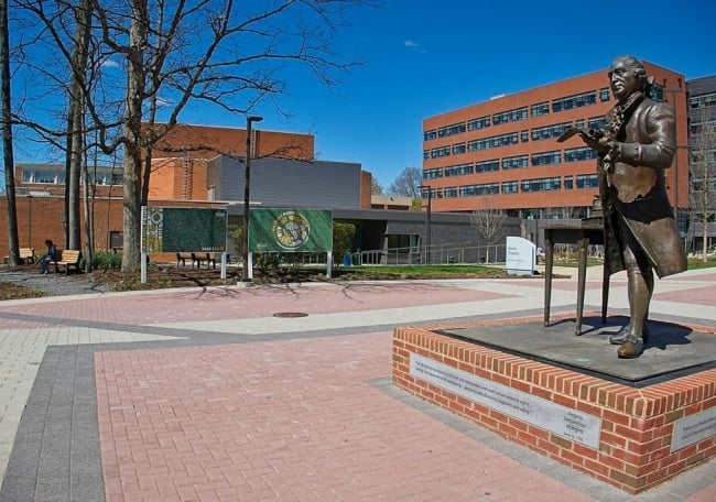 A statue of George Mason sits at the forefront of this image of George Mason University's campus. University buildings can be seen in the background.